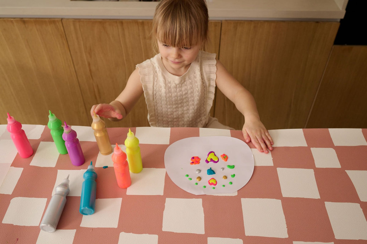 6. Girl painting on a pink and beige checkered table cover with colorful paint bottles