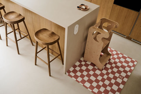 1. Kids mealtime floor mat in pink and beige checkered pattern under a wooden high chair in a modern kitchen setting