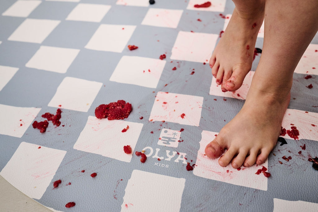 4. Child's feet on blue and white checkered floor mat with food spills highlighting waterproof feature