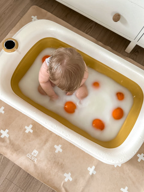 4. Baby playing in tub on beige OLYA KIDS floor mat with cross pattern, surrounded by orange toys