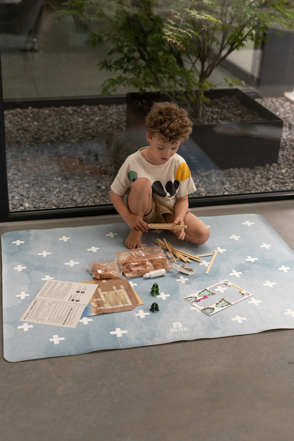 6. Child sitting on blue OLYA KIDS floor mat with white cross pattern, surrounded by craft items, indoors