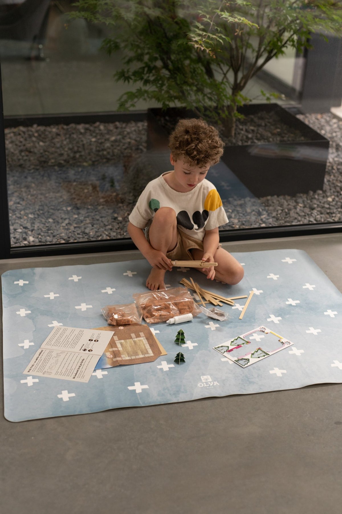 6. Child sitting on blue OLYA KIDS floor mat with white cross pattern, surrounded by craft items, indoors