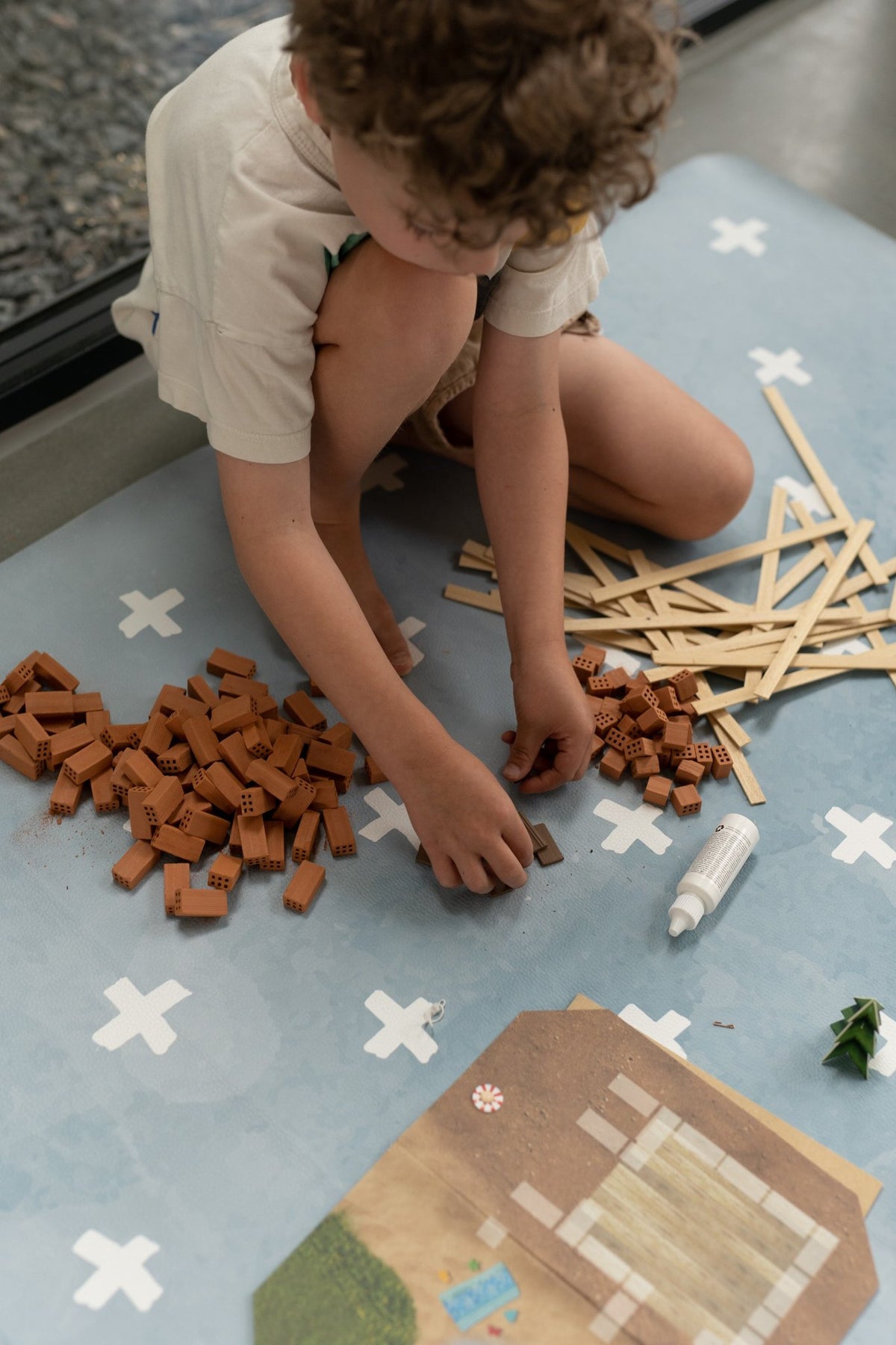 3. Child assembling blocks on blue OLYA KIDS floor mat with white cross pattern, indoors