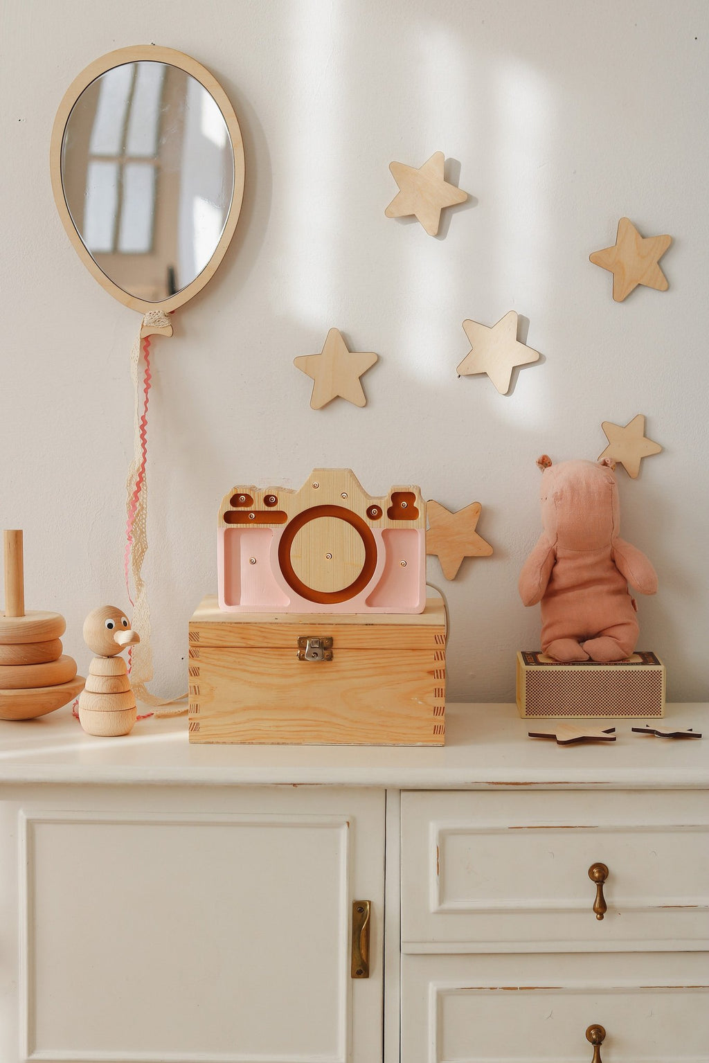 1. Kids room with balloon-shaped mirror on wall, surrounded by wooden star decorations, pink toy camera, and plush toys on white cabinet