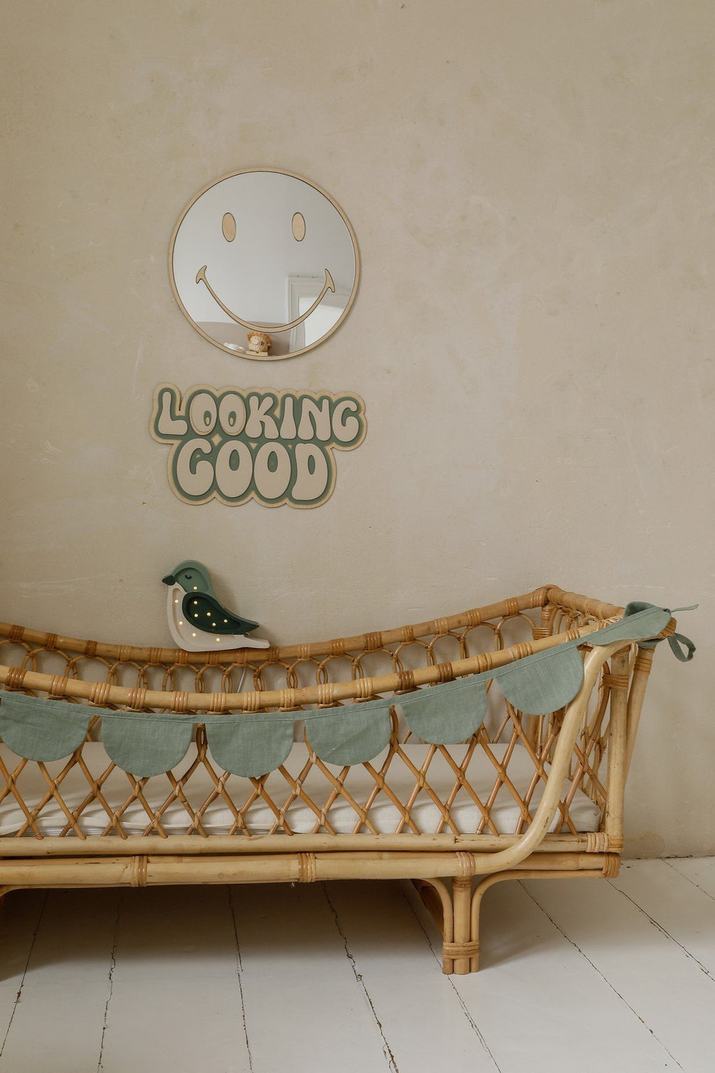 4. Emoji-shaped mirror with gold-tinted acrylic above rattan crib in nursery, featuring 'Looking Good' sign and bird decoration
