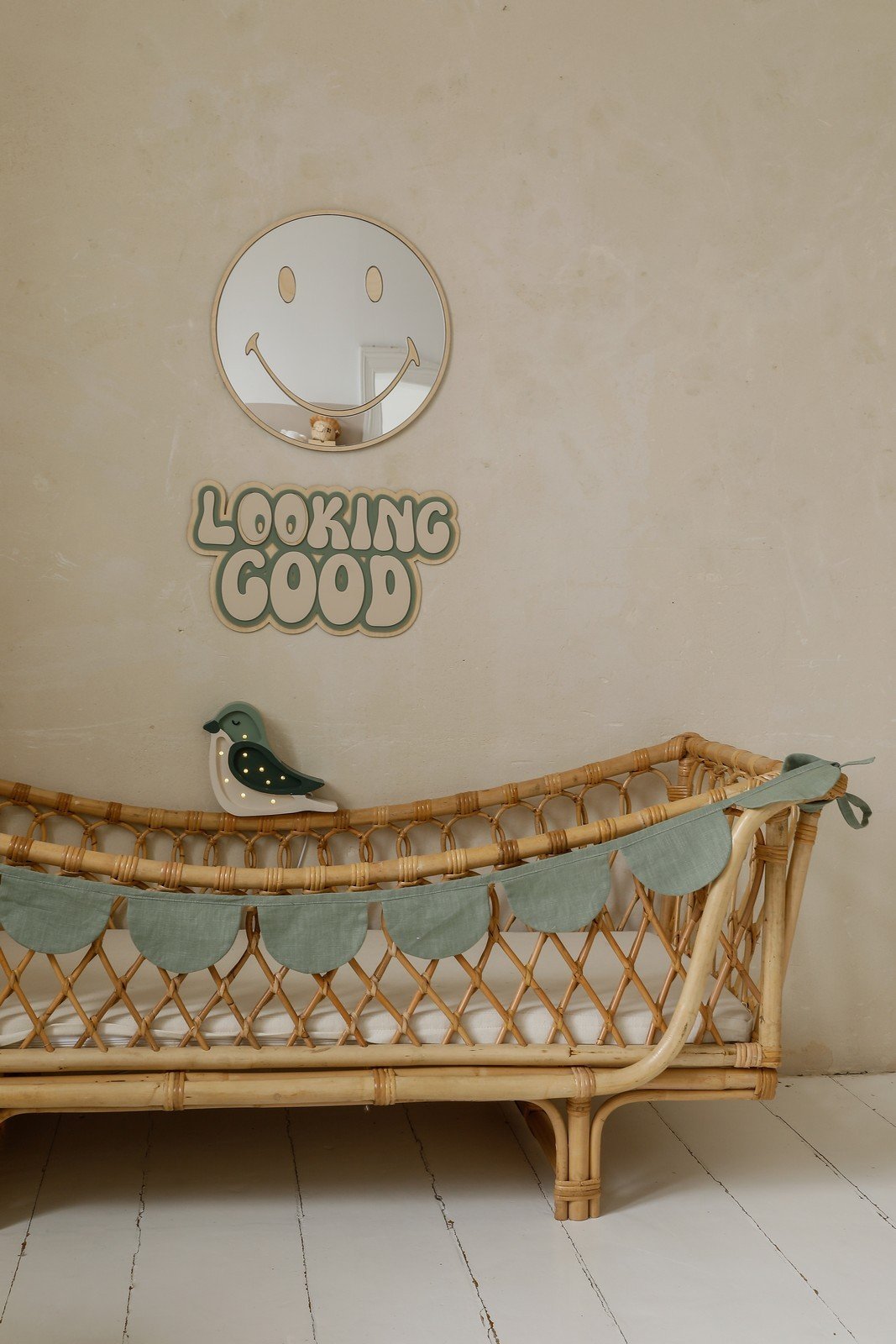 4. Emoji-shaped mirror with gold-tinted acrylic above rattan crib in nursery, featuring 'Looking Good' sign and bird decoration