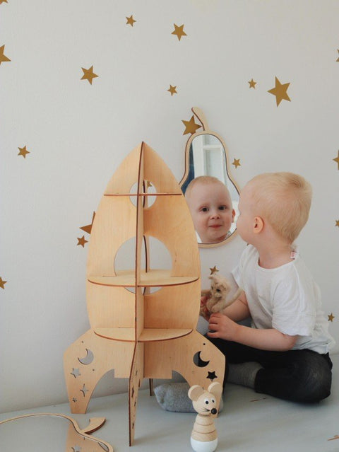 3. Child looking into pear-shaped mirror with plywood frame, surrounded by gold star decals and wooden rocket decor.