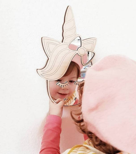 1. Child looking into unicorn-shaped mirror with natural plywood frame, wearing pink hat and sweater, against white wall