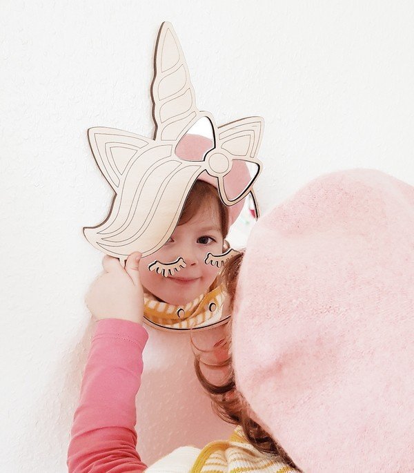 4. Child smiling into unicorn-shaped mirror with natural plywood frame, wearing pink hat and sweater, against white wall