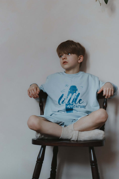 1. Child in pastel blue Zezuzulla shorts and sweatshirt with 'Little Adventure' print sitting cross-legged on a chair