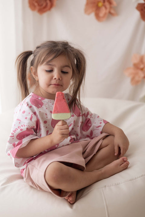3. Young girl sitting cross-legged wearing pastel pink shorts and white printed shirt holding a popsicle in a cozy setting