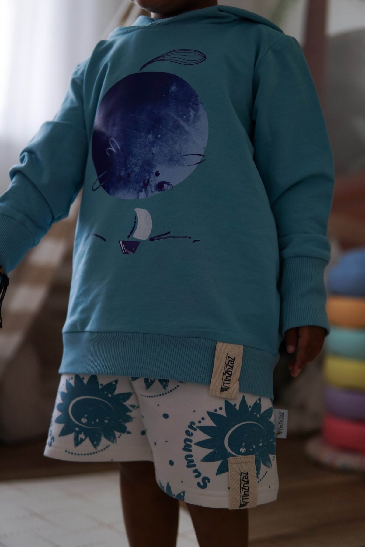 3. Close-up of child wearing white shorts with blue sun print and blue hoodie indoors