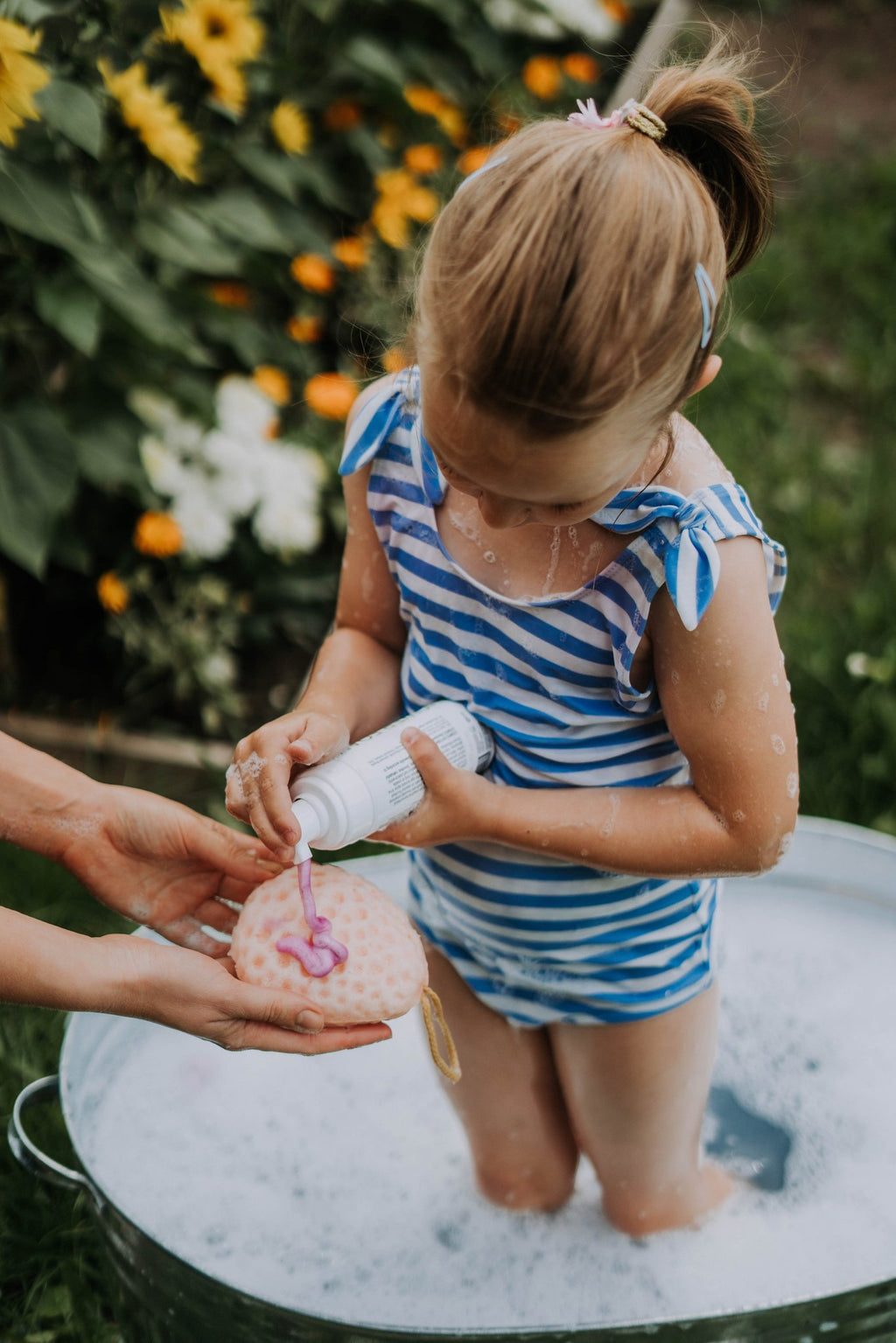 2. Girl in blue striped swimsuit using LUUV Kid's Shower Foam Pear COSMOS with adult assistance in garden