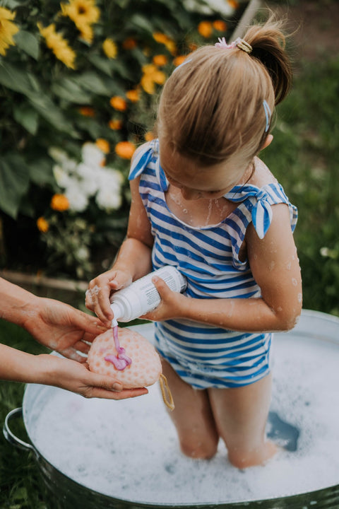 2. Girl in blue striped swimsuit using LUUV Kid's Shower Foam Pear COSMOS with adult assistance in garden