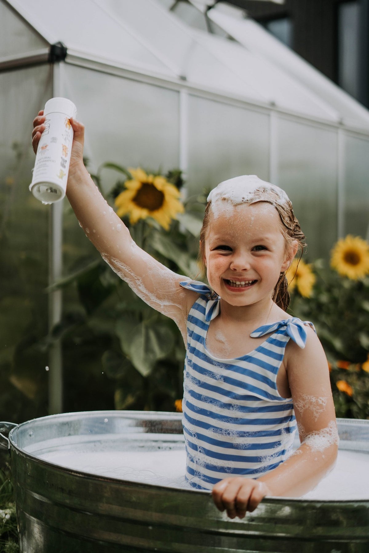 4. Smiling child in blue striped swimsuit holding LUUV Kid's Shower Foam Pear COSMOS in garden with sunflowers