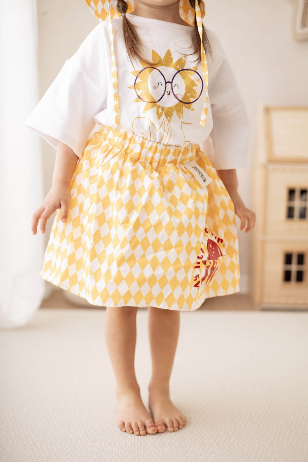 1. Child wearing yellow and white diamond-patterned skirt with red turtle print, paired with a white t-shirt featuring a sun graphic, standing in a cozy room