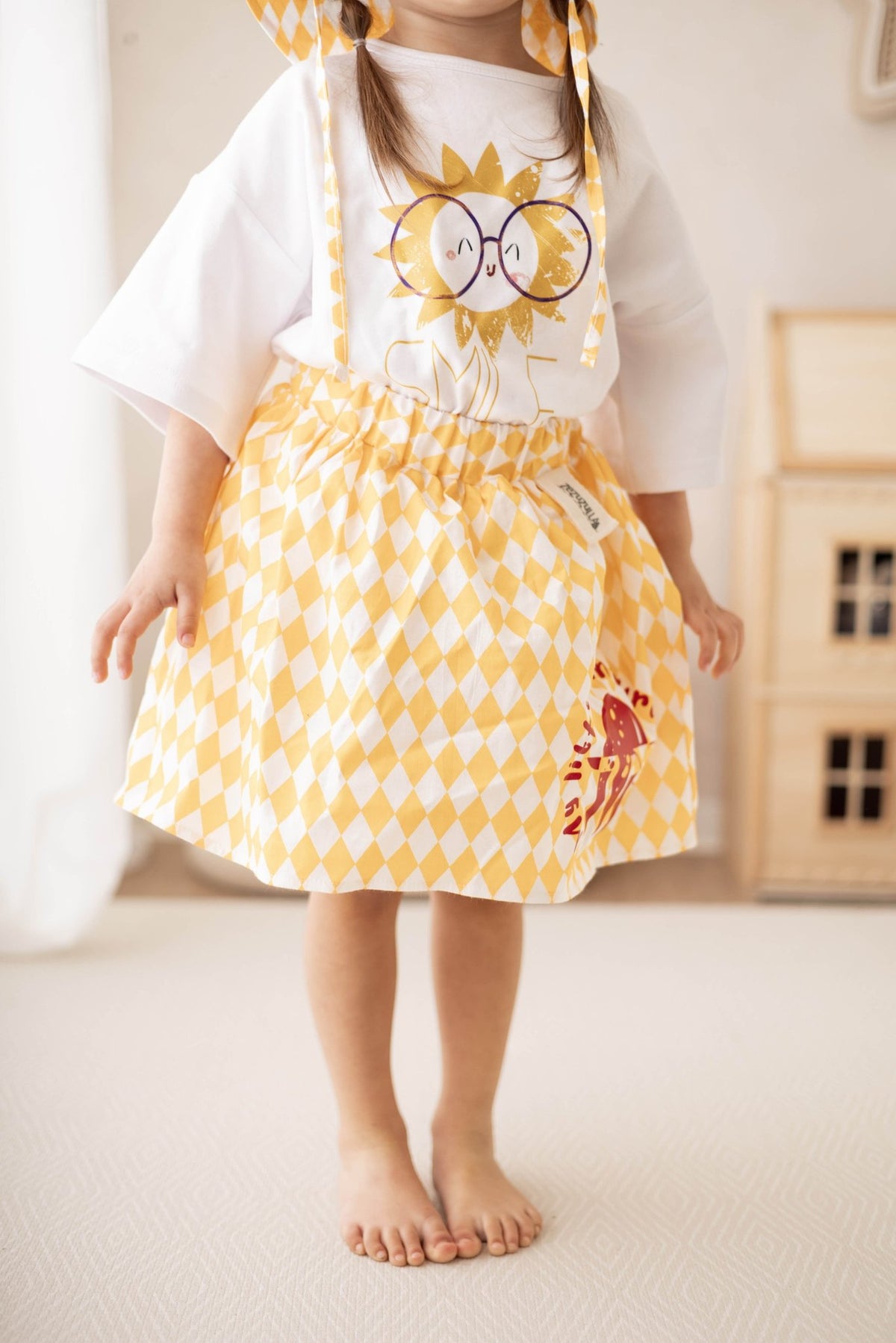 1. Child wearing yellow and white diamond-patterned skirt with red turtle print, paired with a white t-shirt featuring a sun graphic, standing in a cozy room
