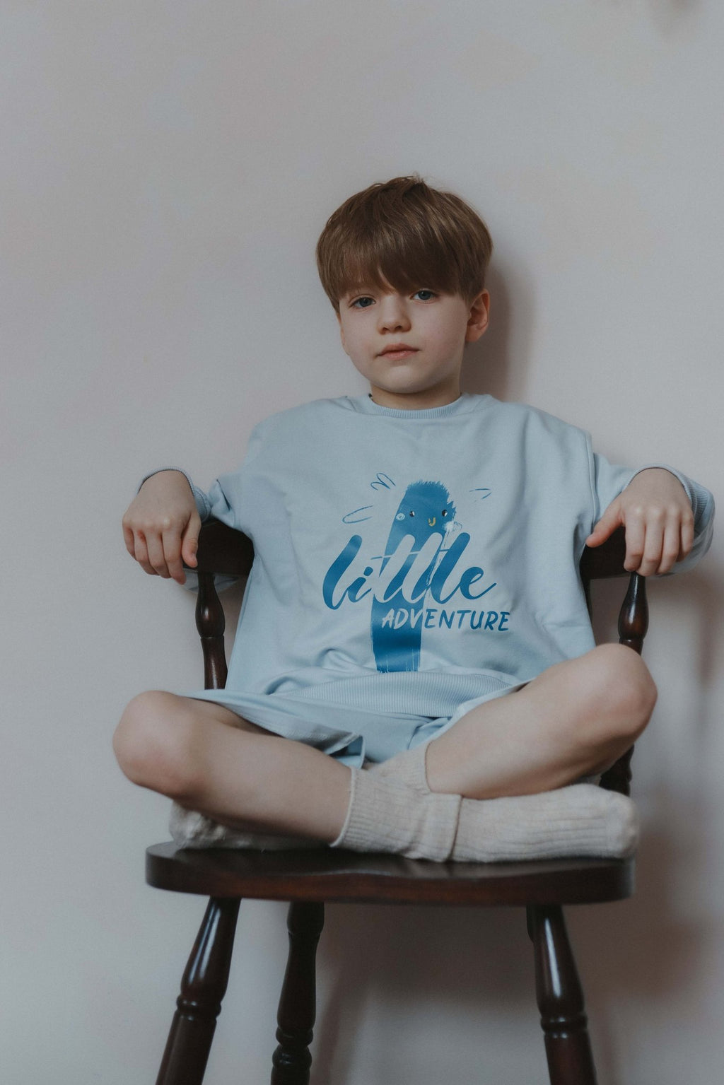 1. Boy wearing light blue Zezuzulla sweatshirt with 'Little Adventure' print sitting on a chair