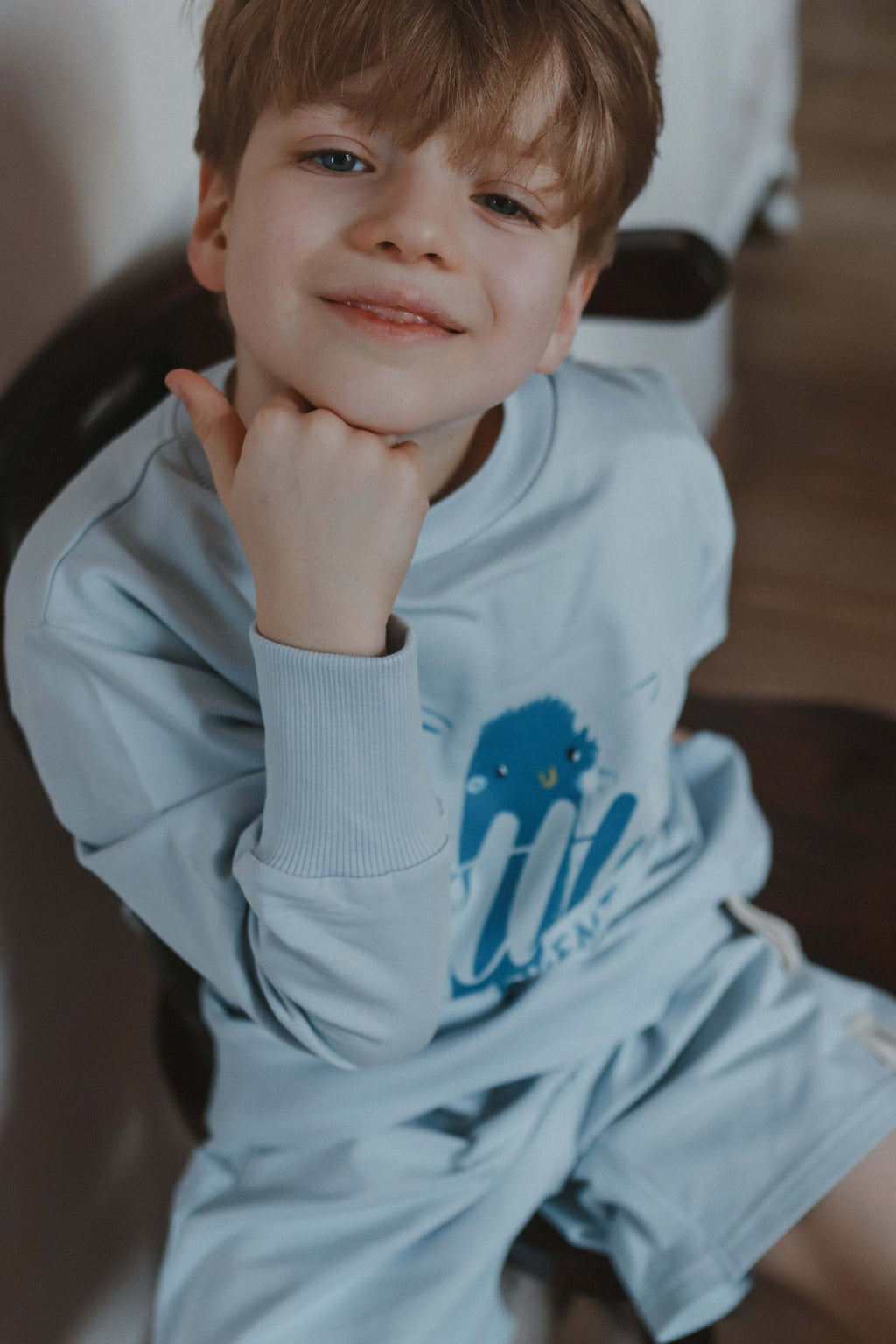 1. Smiling boy in light blue Zezuzulla sweatshirt with 'Little Adventure' print sitting on chair