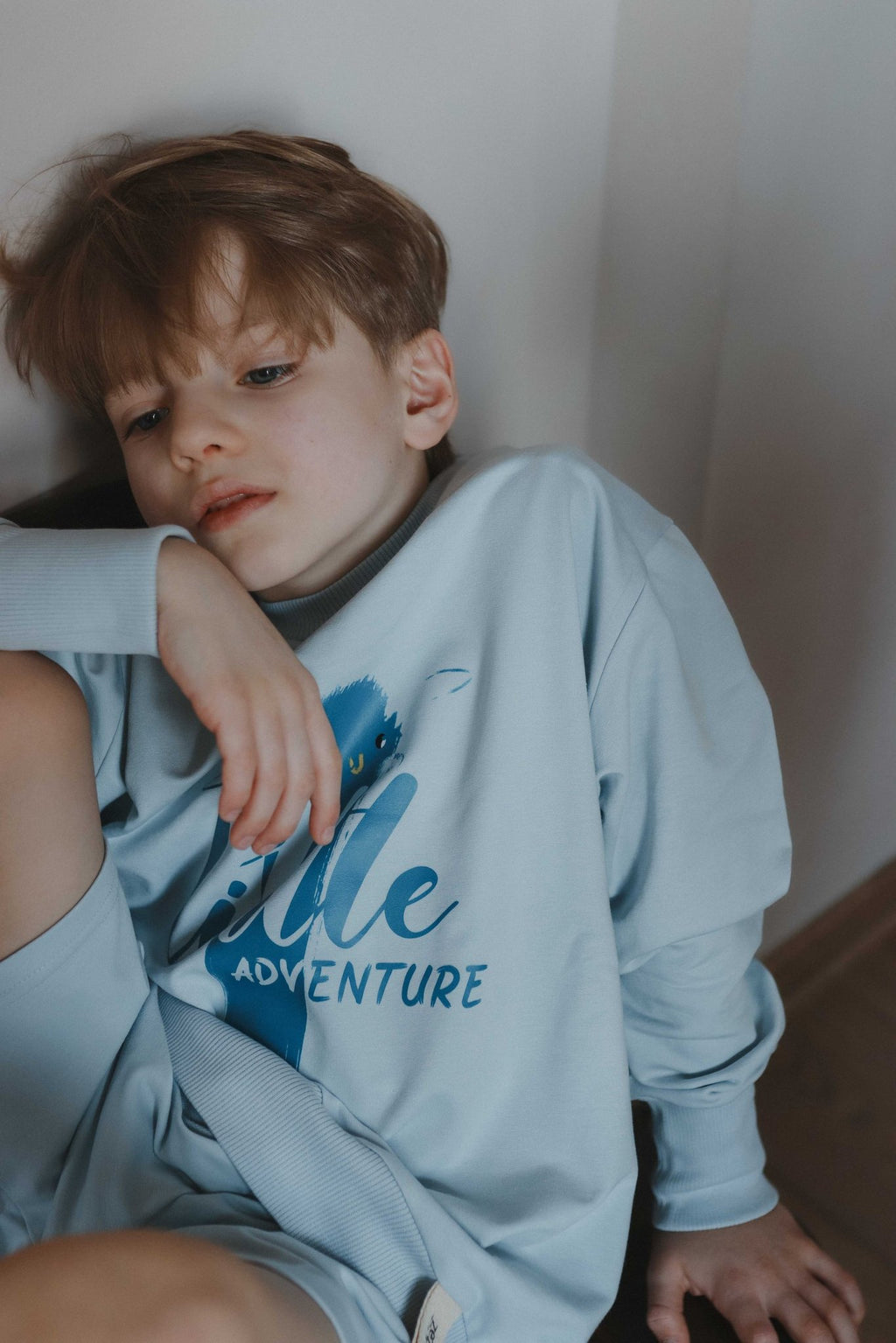 1. Boy in light blue Zezuzulla sweatshirt with 'Little Adventure' print leaning against wall
