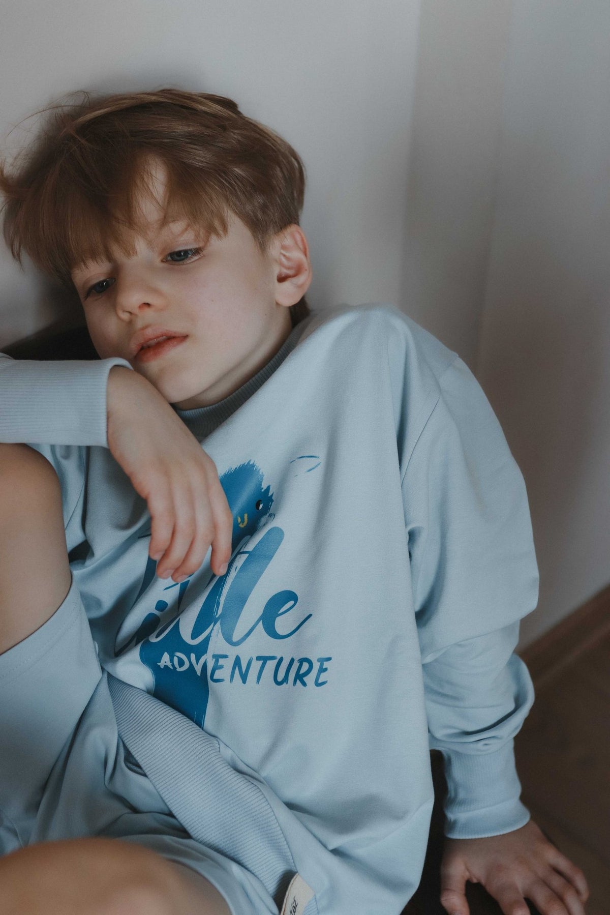 1. Boy in light blue Zezuzulla sweatshirt with 'Little Adventure' print leaning against wall
