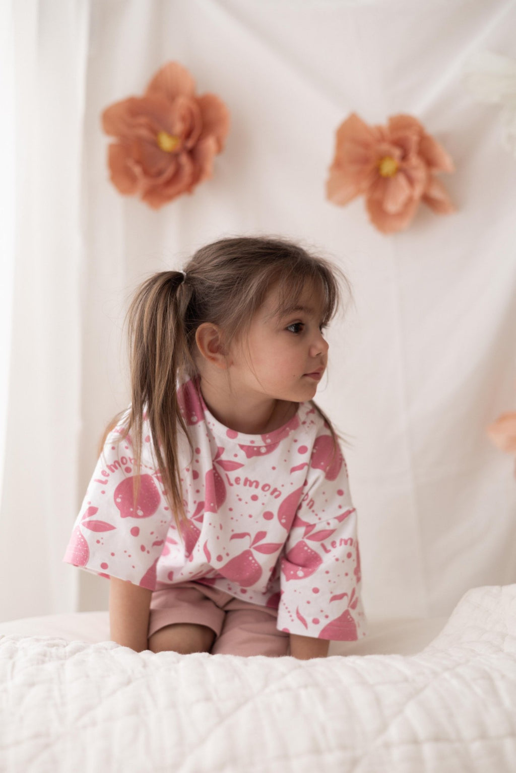 1. Young girl wearing Zezuzulla white T-shirt with pink lemon print, sitting on a bed with floral backdrop