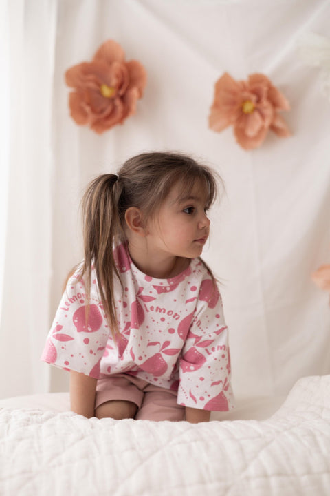 1. Young girl wearing Zezuzulla white T-shirt with pink lemon print, sitting on a bed with floral backdrop
