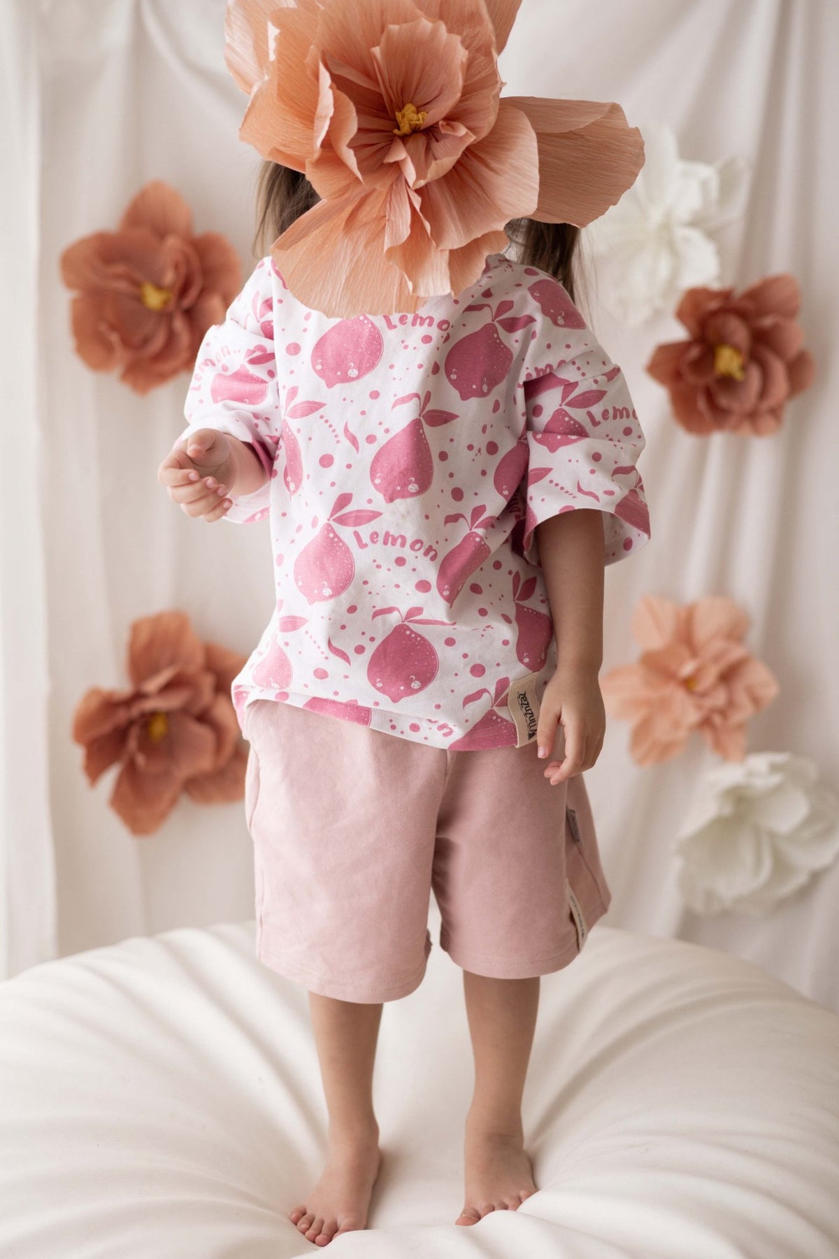 2. Child standing with back to camera in Zezuzulla white T-shirt with pink lemons, surrounded by large paper flowers