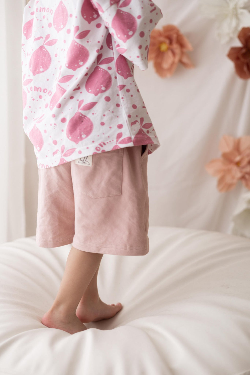 3. Close-up of child wearing Zezuzulla T-shirt with pink lemon print and pink shorts, standing on white cushion