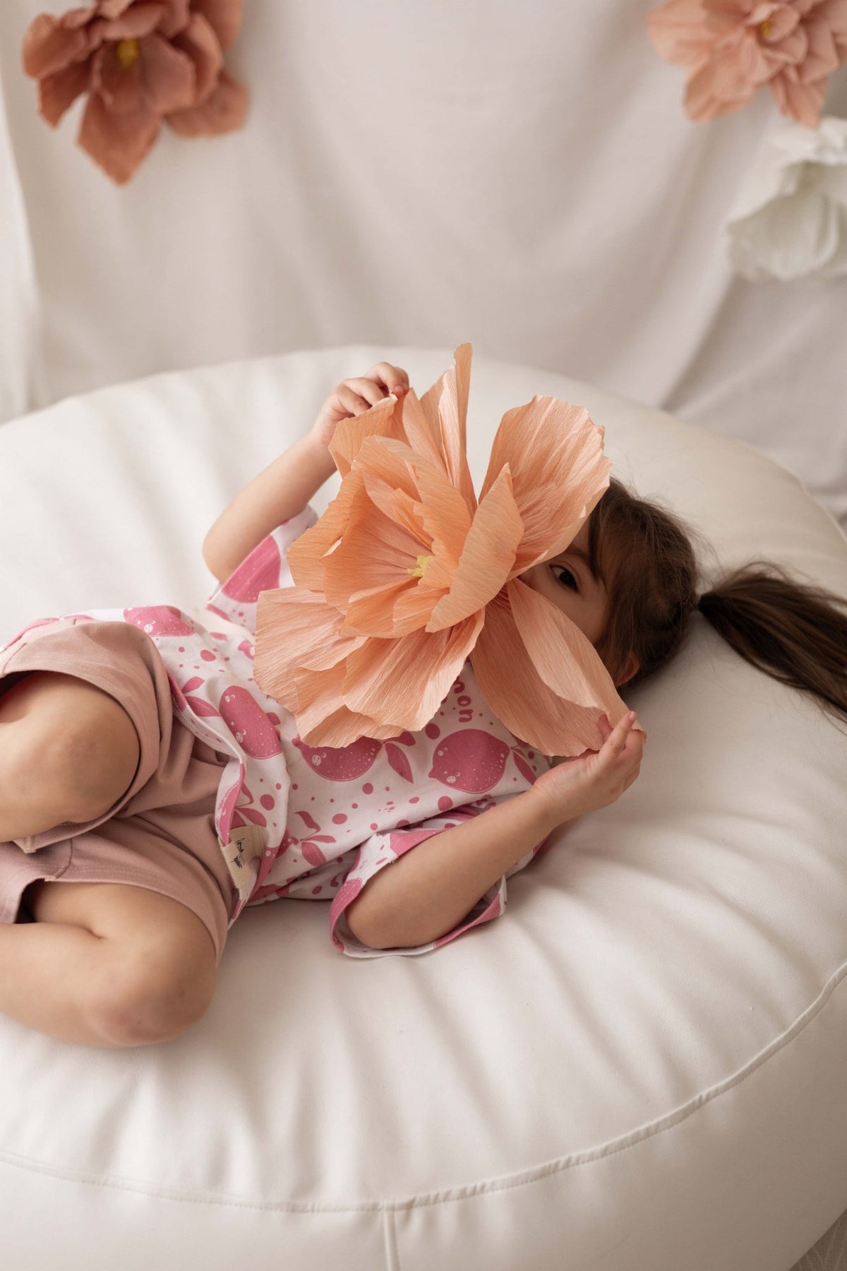 3. Child lying on white cushion wearing Zezuzulla T-shirt with pink lemons, holding large paper flower