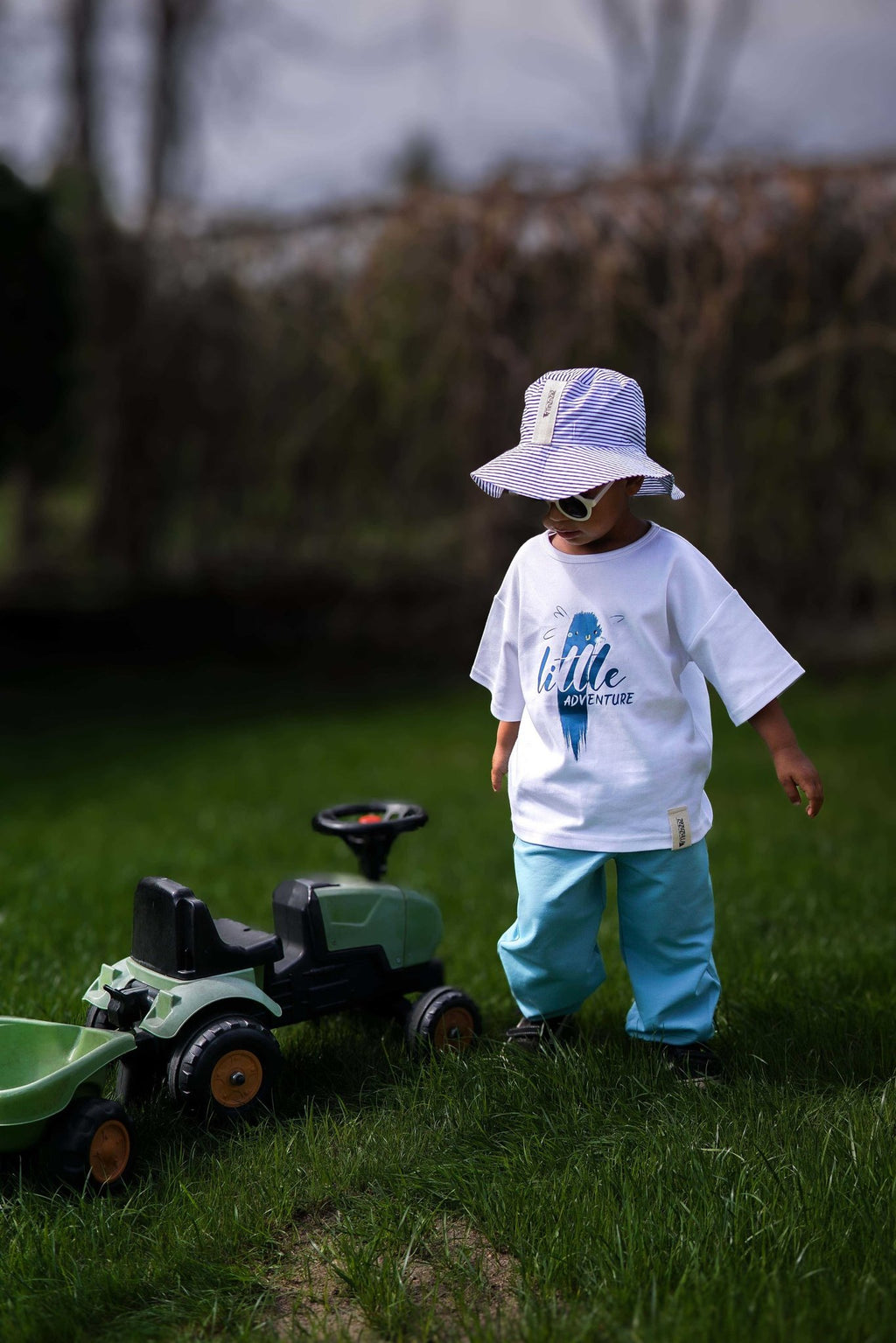 3. Child in white Zezuzulla T-shirt with 'little adventure' print, wearing a hat and sunglasses, standing on grass with a toy tractor