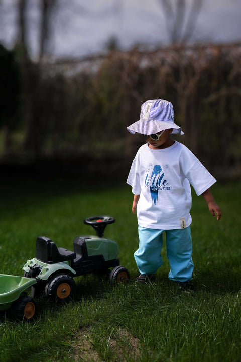 3. Child in white Zezuzulla T-shirt with 'little adventure' print, wearing a hat and sunglasses, standing on grass with a toy tractor
