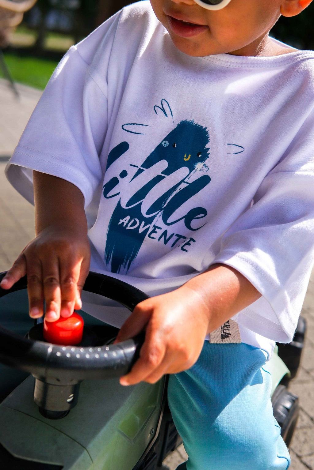 1. Child wearing white Zezuzulla T-shirt with 'little adventure' print, playing on a toy vehicle outdoors