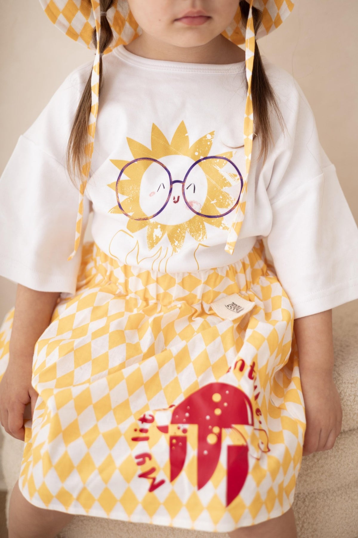 3. Close-up of child sitting, wearing white Zezuzulla T-shirt with sun and 'SMILE' print, paired with yellow checkered skirt
