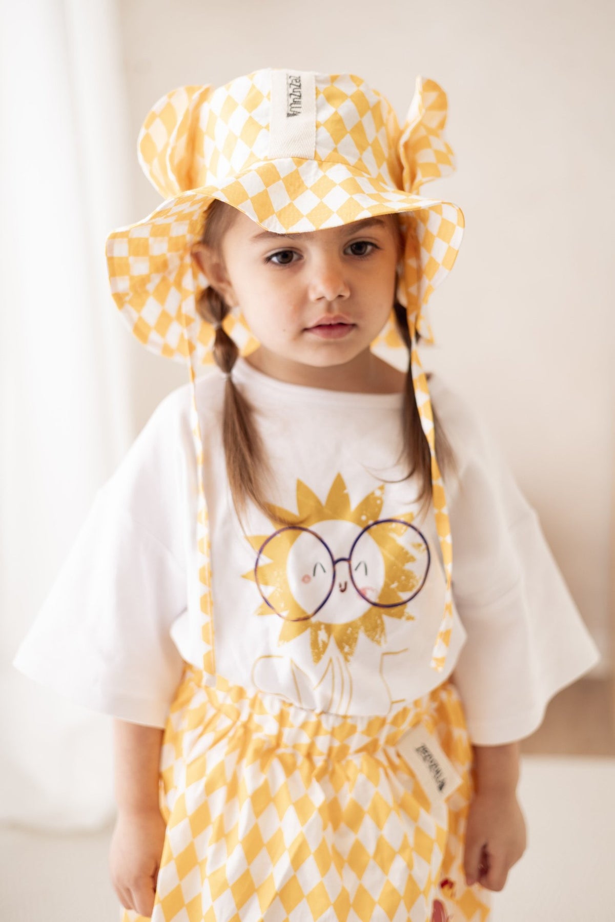 1. Young girl wearing white Zezuzulla T-shirt with sun and 'SMILE' print, paired with yellow checkered hat and skirt, standing indoors