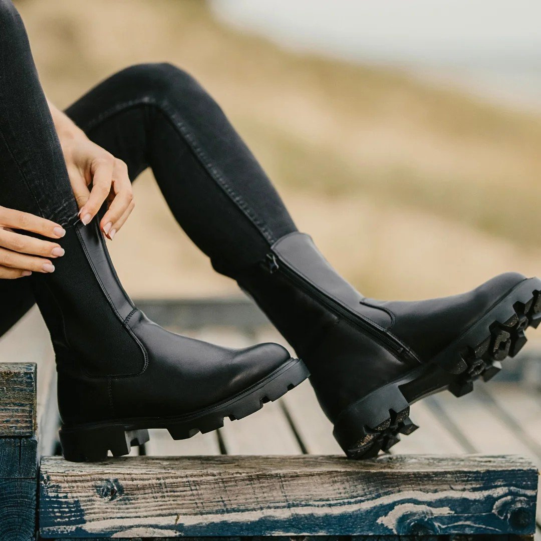 1. Woman wearing black KIRA Chelsea Rebel boots with extended shaft sitting on wooden steps outdoors