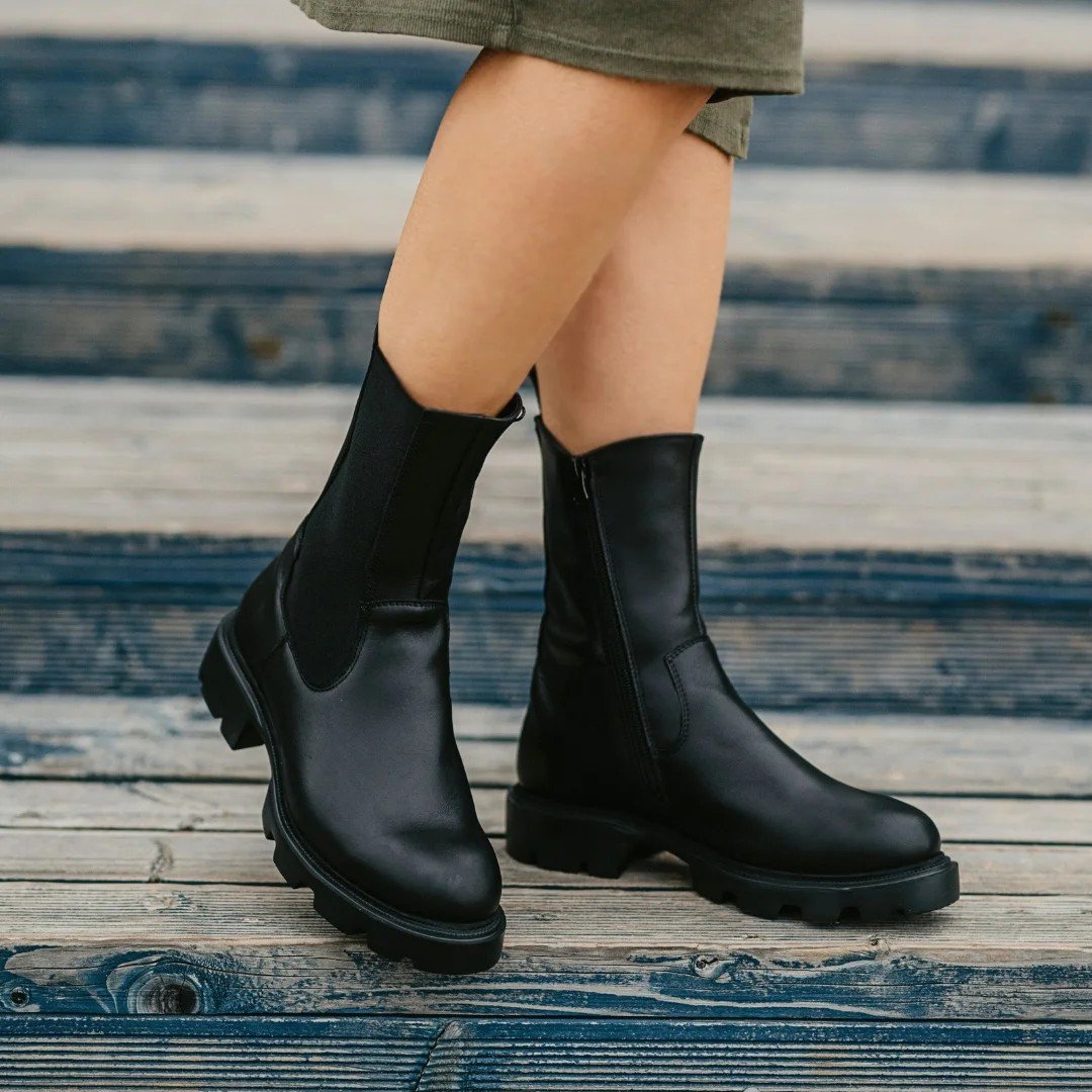 1. Woman wearing black KIRA Chelsea Rebel boots with durable sole standing on wooden planks