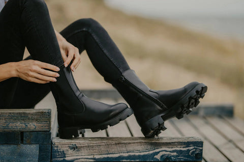 2. Woman in black jeans adjusting black KIRA Chelsea Rebel boots with side zipper on wooden steps