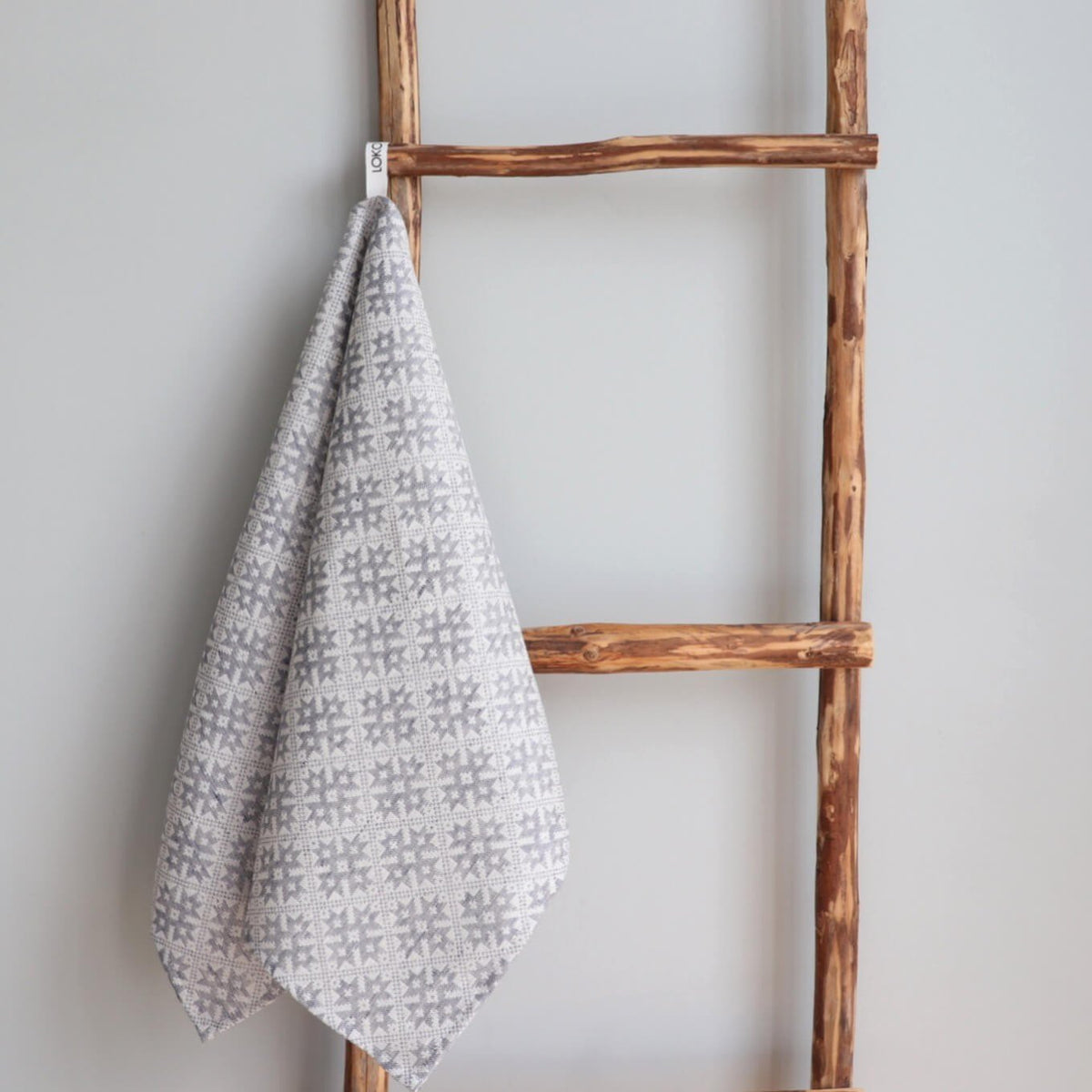 1. Kitchen towel with Pärnumaa folk art pattern hanging on rustic wooden ladder, showcasing grey and white design