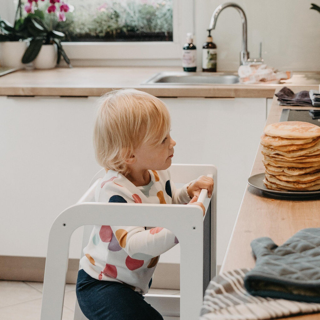3. Child using white Montessori kitchen helper tower in kitchen with stack of pancakes