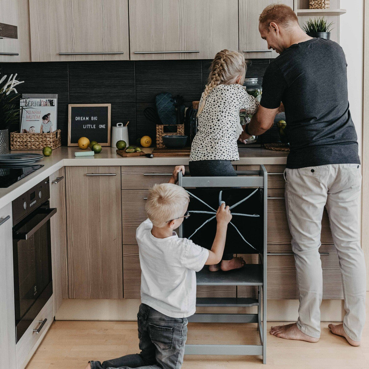 1. Family with children using grey Montessori kitchen helper tower in kitchen, child drawing on blackboard