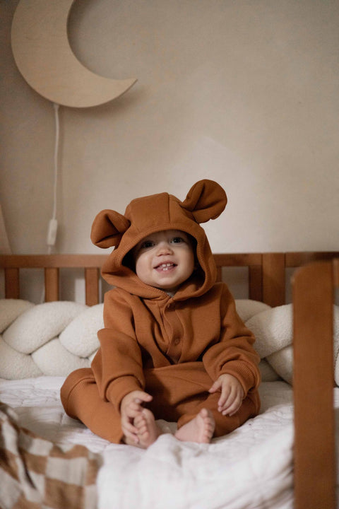 1. Baby in cinnamon eared jumpsuit with hood, sitting on bed with moon decor in background