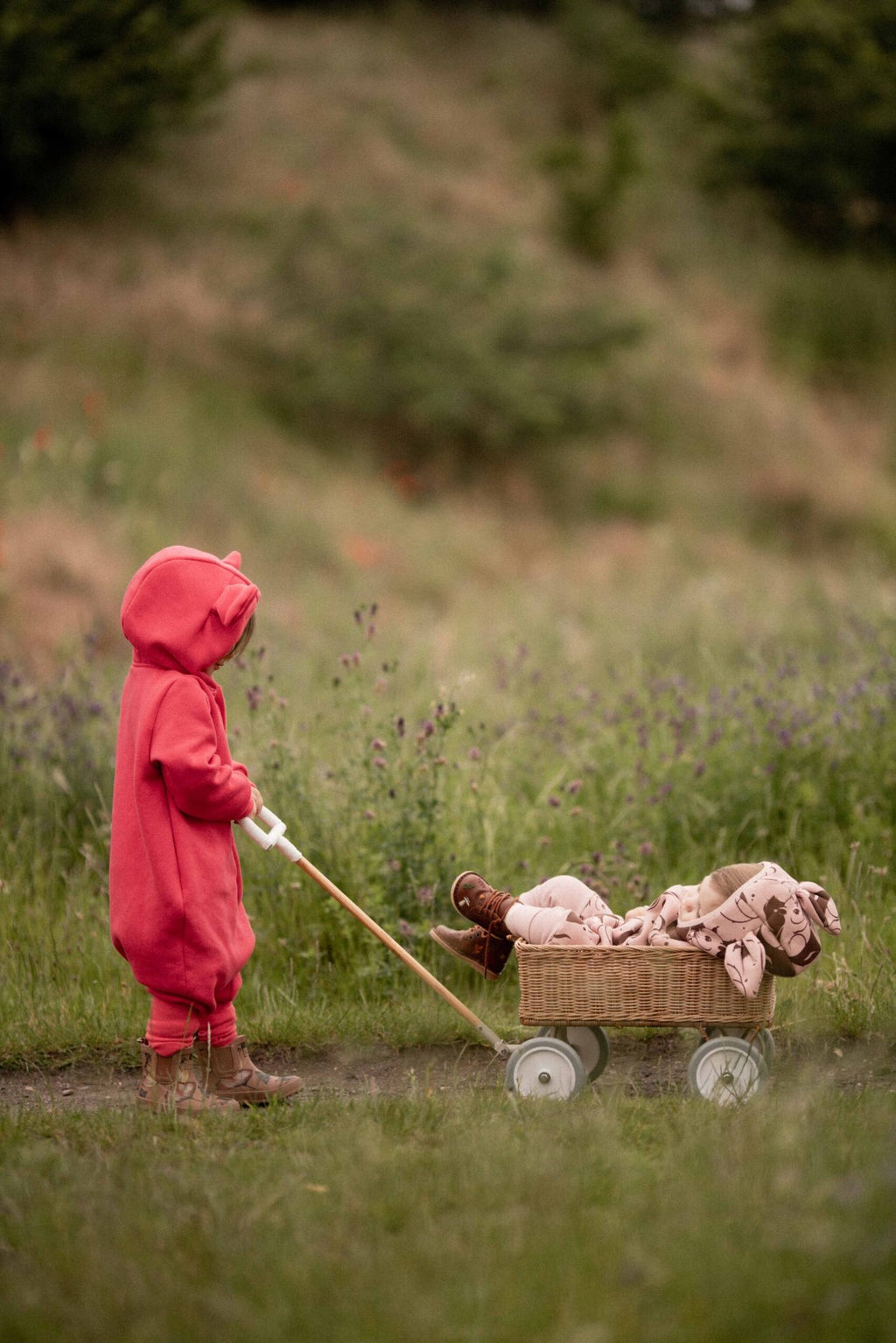 2. Child in coral eared jumpsuit by Zezuzulla pulling a wagon in a grassy field
