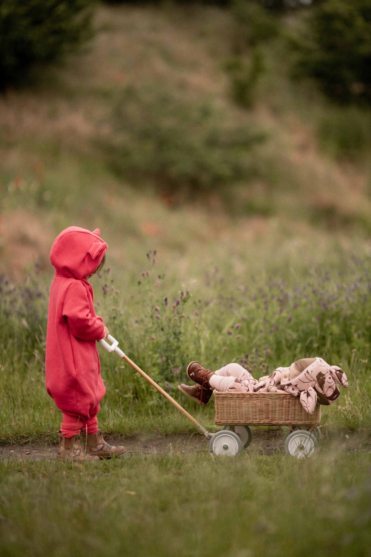 2. Child in coral eared jumpsuit by Zezuzulla pulling a wagon in a grassy field