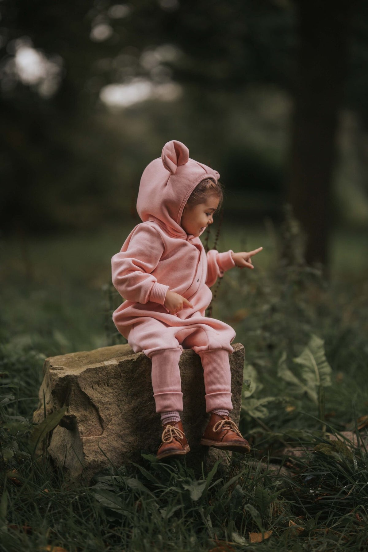 1. Child wearing peach eared jumpsuit sitting on a log in a forest setting
