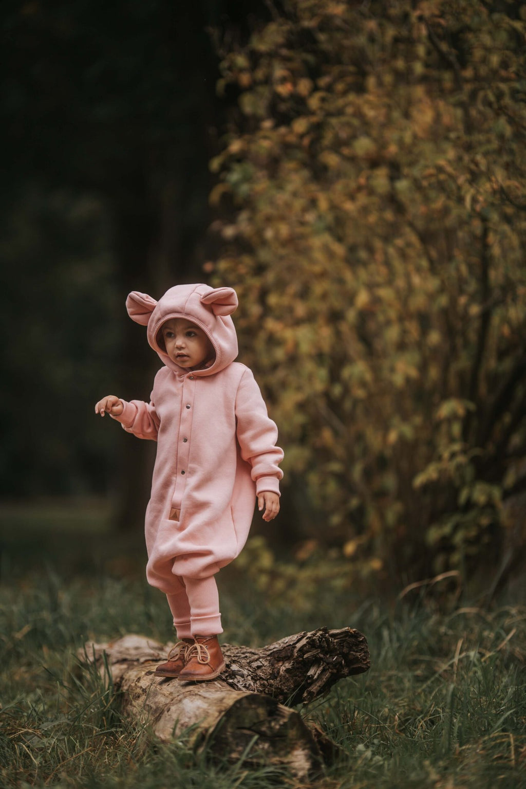 1. Child standing on a log wearing peach eared jumpsuit in a wooded area