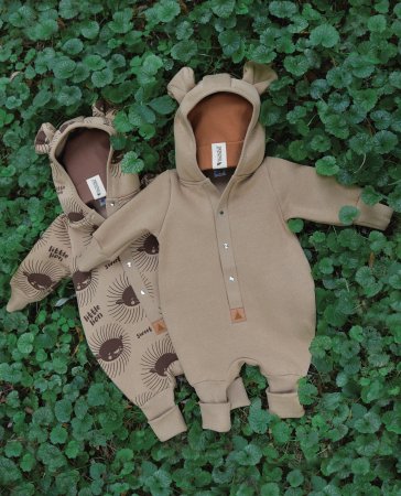 4. Two eared jumpsuits for babies, one in cacao and one with sun print, laid on green foliage