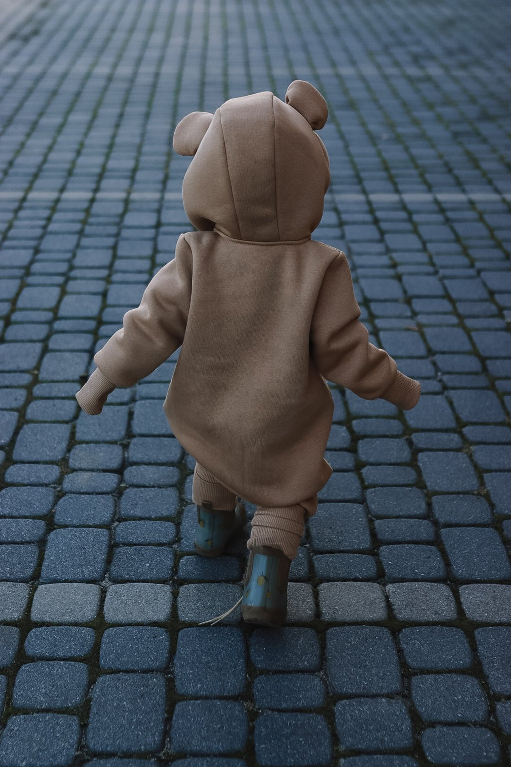 1. Baby wearing cacao eared jumpsuit walking on cobblestone path, showing back view with hood and ears