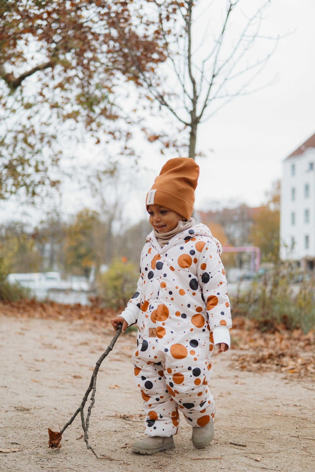 1. Child wearing black and orange polka dot winter jumpsuit with teddy bear ears, walking on a path with autumn leaves