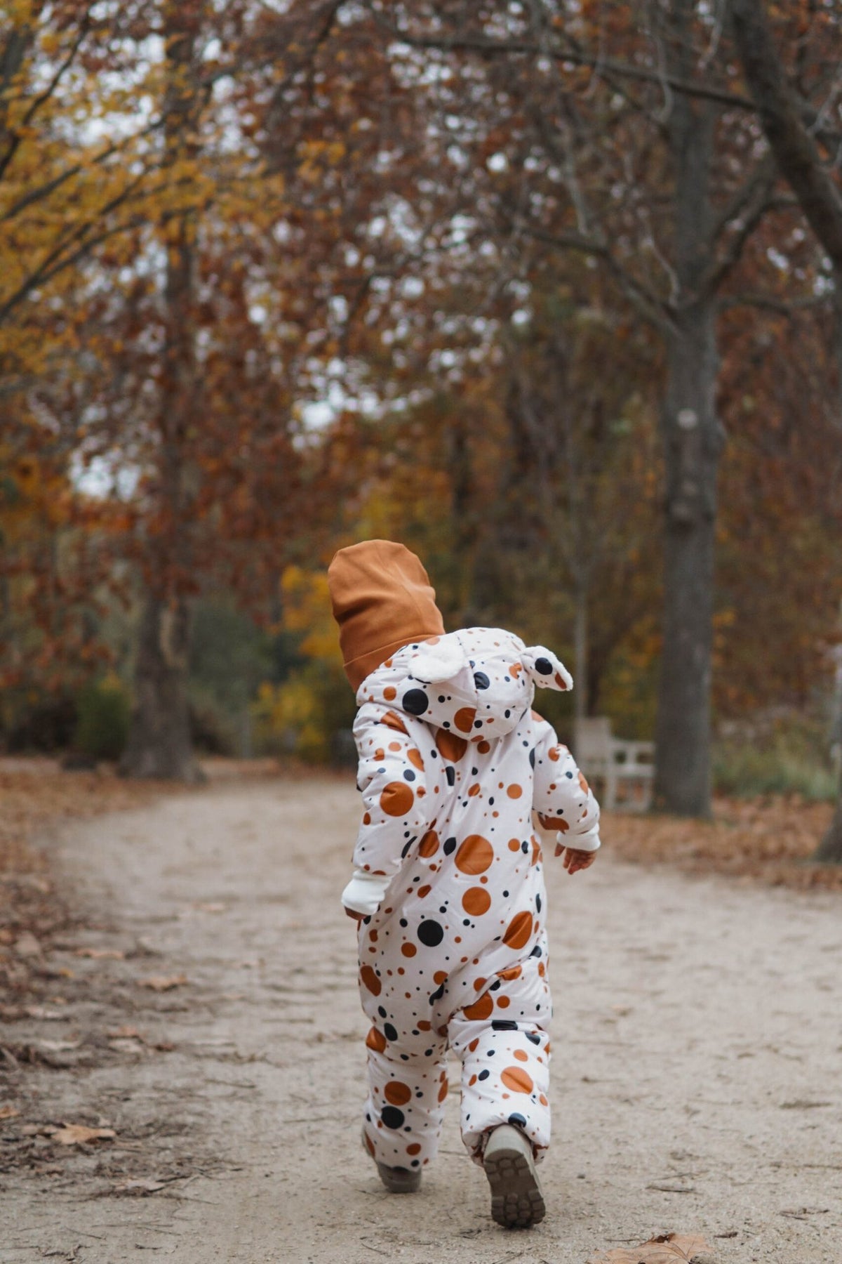 1. Child wearing black and orange polka dot winter jumpsuit with teddy bear ears, walking away on a forest path